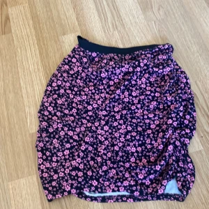 Blommig kjol i rosa och svart - Säljer en söt blommig kjol med rosa blommor på svart bakgrund. Kjolen har en tight passform och är perfekt för vår och sommar. Den är knälång och har en elastisk midja för extra komfort.