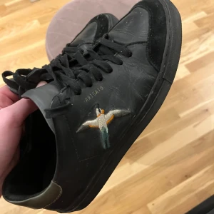 Svarta sneakers från Axel Arigato - Snygga svarta sneakers från Axel Arigato med broderad fågel på sidan. Skorna har snörning och en rund tå. Perfekta för en stilren look.