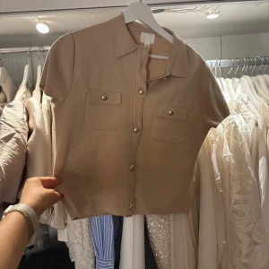 Beige kortärmad kofta med knappar - Säljer en stilren beige kofta med korta ärmar och dekorativa guldknappar. Koftan har två fickor framtill och en klassisk krage. Perfekt för en elegant look.