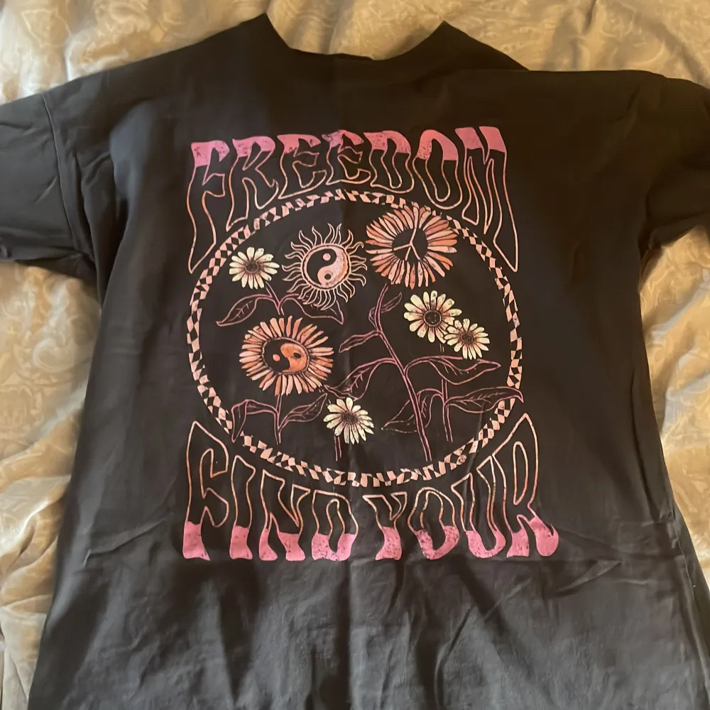 Säljer en grå t-shirt med ett coolt tryck i rosa och vitt. Texten 'FREEDOM FIND YOUR' omger ett motiv av blommor och ett yin-yang-tecken. Perfekt för en avslappnad stil.. T-paidat.