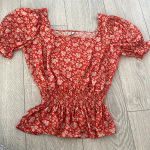 Röd blommig blus - Charmig röd blommig blus med korta puffärmar och smockdetaljer i midjan. Perfekt för en somrig look med sitt lätta och luftiga material.