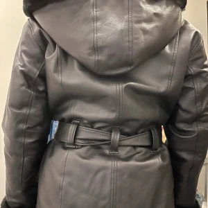 Svart skinnjacka från Wilsons Leather - Snygg svart skinnjacka från Wilsons Leather med mjukt foder och huva. Jackan har dragkedja och bälte i midjan för en justerbar passform. Perfekt för kyliga dagar med stil.🧚🏽🌸