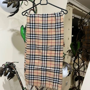 Rutig BURBERRY halsduk - Snygg rutig halsduk i beige med svarta och röda ränder. Gjord av en blandning av pashmina och silke, vilket ger en mjuk och lyxig känsla. Perfekt för att hålla sig varm och stilren under kyliga dagar.Det ni ser på bilden är det ni får😁 ett plagg som är köpt i bulk så jag vet lila lite som ni, men jag har kollat så gott jag kan på alla möjliga sajter och det jag kan kolla stämmer överens med originalet väldigt bra tröja raka o fina sömmar, kvalitets tyg