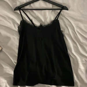Svart linne med spetsdetaljer - Snyggt svart linne med tunna axelband och vacker spetsdetalj vid halsringningen. Perfekt för en elegant look. Är omsydd i ryggen för en fina passform 