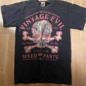 Svart t-shirt med tryck från Fruit of the Loom - Cool svart t-shirt från Fruit of the Loom med ett stort tryck av en dödskalle och texten 'Vintage Evil' i rött. Perfekt för dig som gillar en edgy stil. Tillverkad i tung bomull för en skön känsla.