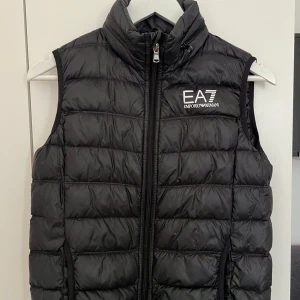 Svart dunväst från Emporio Armani - Snygg svart dunväst från Emporio Armani med dragkedja och logga på bröstet. Perfekt för kyligare dagar med sin quiltade design. Västen har en hög krage för extra värme och stil.