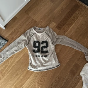 Vit och beige tröja från FB Sister - Säljer en cool vit och beige tröja från FB Sister med texten '92 Future Legend' på framsidan. Tröjan har långa ärmar med ett nätliknande mönster och rund halsringning. Perfekt för en avslappnad stil!