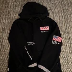 Svart hoodie  - Säljer en svart hoodie med NASA-logga och amerikansk flagga på ärmen. Tröjan har en stor ficka framtill och textdetaljer på ärmen och ryggen. Köpt på hm i storlek L