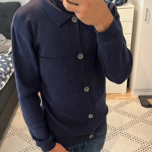 Mörkblå kofta med knappar - Säljer en stilren mörkblå kofta med knappar framtill. Koftan har en klassisk krage och en bröstficka. Perfekt för en avslappnad men ändå uppklädd look.