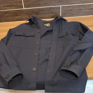 Mörkblå overshirt  - Säljer en stilren mörkblå overshirt från H&M i regular fit. Jackan har knappar framtill och två bröstfickor med lock. Perfekt för lager-på-lager-stil och passar till många olika outfits.