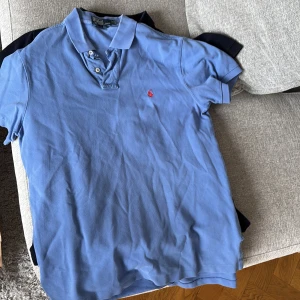 Blå pikétröja från Ralph Lauren - Säljer en klassisk blå pikétröja från Ralph Lauren med korta ärmar och en liten röd logotyp på bröstet. Perfekt för en stilren och avslappnad look. Tröjan har knappar vid kragen för en justerbar passform.