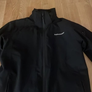 Svart jacka från Peak Performance - Säljer en stilren svart jacka från Peak Performance. Jackan har en dragkedja framtill och en hög krage för extra skydd. Perfekt för kyligare dagar med sin funktionella design och diskreta logga på bröstet.