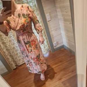 Blommig klänning från Vintage Stories - Härlig blommig sommarklänning från Vintage Stories i en ljus rosa nyans. Klänningen har trekvartsärmar och ett vackert blommönster . Den är v-ringad och har en omlott-design. Väldigt skrynklig på bilden.