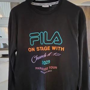Svart långärmad tröja från Fila - Säljer en svart långärmad tröja från Fila med färgglad text på framsidan och baksidan. Texten inkluderar 'On Stage With' och 'World Tour' i orange, samt andra detaljer i turkos, lila och rosa. Perfekt för dig som gillar en unik och trendig stil.