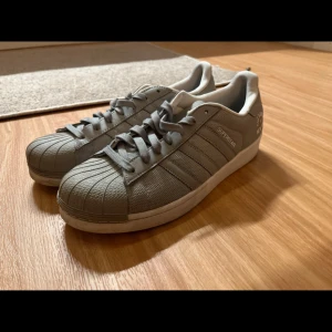 Grå Adidas Superstar sneakers - Snygga grå Adidas Superstar sneakers med klassisk snäckformad tå och snörning. Skorna har de ikoniska tre ränderna på sidan och en vit sula. Perfekta för en stilren look.