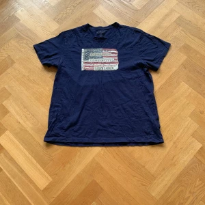 Mörkblå t-shirt från Ralph Lauren - Säljer en mörkblå t-shirt från Ralph Lauren med ett coolt tryck av en amerikansk flagga på bröstet. Är ganska liten i storleken. XL men passar även L. Moddellen är 184 och väger 70 kg. Priset kan diskuteras.
