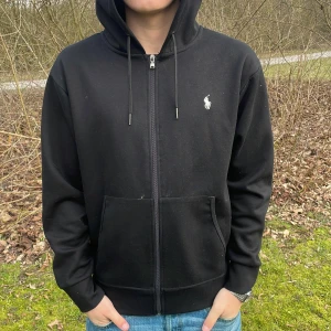 Svart hoodie från Polo Ralph Lauren - Säljer en stilren svart hoodie från Polo Ralph Lauren med dragkedja och huva. Den har en liten logotyp på bröstet och är tillverkad i ett mjukt material. Perfekt för en avslappnad look. den är köpt begagnat så äktheten kan inte bevisas