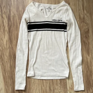 Vit långärmad tröja från Hollister - Säljer en vit långärmad tröja från Hollister med svarta ränder över bröstet och en liten logga. Tröjan har en v-ringad hals och är ribbad i materialet. Perfekt för en avslappnad stil. Och är figurnära