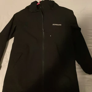 Svart regnjacka från Didriksons - Säljer en svart regnjacka från Didriksons, perfekt för regniga dagar. Jackan är 100% vattentät och har en dragkedja framtill. Den har långa ärmar och en praktisk huva för extra skydd mot väder och vind.