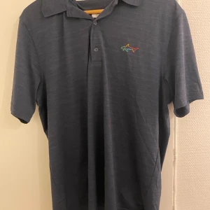 Golf piké marinblå - Snygg mörkblå pikétröja med korta ärmar och en färgglad broderad logga på bröstet. Perfekt för en avslappnad stil. Tröjan har en klassisk krage och knappar framtill. Golf piké 