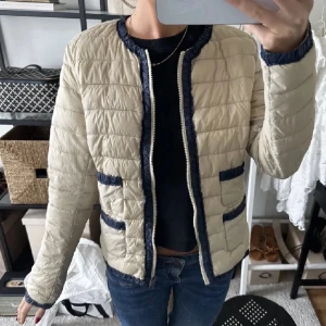 Beige quiltad jacka med blå detaljer - Snygg beige quiltad jacka med blå detaljer och dragkedja framtill. Jackan har långa ärmar och två fickor med blå kant. Perfekt för en stilren look.