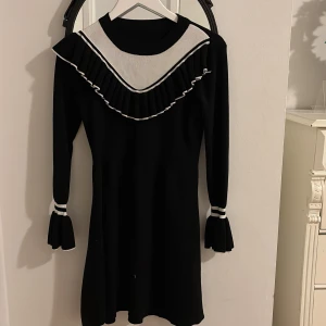 Svart klänning med vit detalj - Elegant svart klänning med vita detaljer och volang vid kragen. Långärmad design med dekorativa vita ränder vid ärmslut. Perfekt för en stilren look.