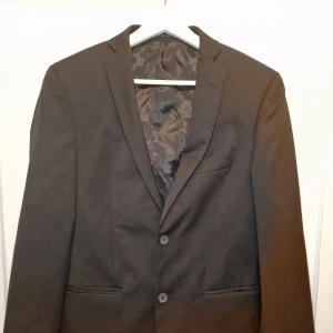 Svart kavaj  - Snygg svart kavaj i slim fit. Kavajen har två knappar framtill och en klassisk krage. Insidan är fodrad med ett elegant mönster och har röda detaljer. 