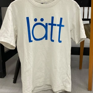 Beige t-shirt med blå text - Säljer en beige t-shirt med blå text 'lätt' tryckt på framsidan. T-shirten har en rund hals och korta ärmar. Perfekt för en avslappnad stil. Storleken är S men känns mer som M.