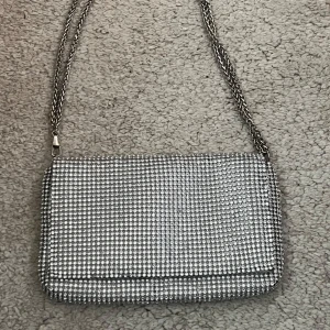 Silverfärgad axelväska med kedja - Snygg silverfärgad axelväska med glittrigt mönster och kedjeaxelrem. Väskan har en praktisk magnetknapp för enkel öppning och stängning. Perfekt för att ge en extra touch till din outfit.