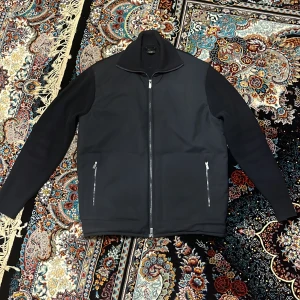 Svart jacka från Massimo Dutti - Snygg svart jacka från Massimo Dutti med dragkedja framtill och två fickor med dragkedjor. Jackan har en stilren design med ribbade detaljer på kragen och ärmarna. Perfekt för en stilren look.