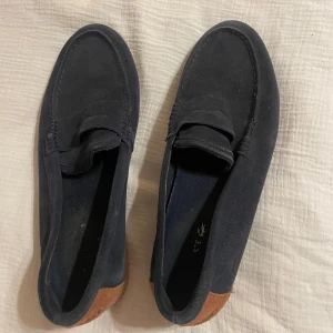 Mörkblå loafers från lacoste - Snygga mörkblå loafers från Lacoste med en stilren design och brun detalj vid hälen. Perfekta för en avslappnad men ändå elegant look. Skorna har en klassisk form och är tillverkade i mocka för en lyxig känsla. Nypris 1400