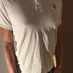 Vit pikétröja från Moncler - Snygg vit pikétröja från Moncler med klassisk krage och knappar. Tröjan har en liten logotyp på bröstet och ränder i rött och blått på axlarna. Perfekt för en stilren look.