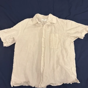 Beige linneskjorta från Uniqlo - Säljer en stilren beige kortärmad skjorta från Uniqlo i linne. Skjortan har en klassisk krage och knappar framtill samt en bröstficka. Perfekt för varma dagar!