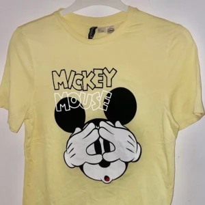 Gul Mickey Mouse t-shirt - Säljer en gul t-shirt med ett stort tryck av Mickey Mouse som täcker ögonen. Perfekt för alla Disney-fans! T-shirten har en rund hals och korta ärmar.