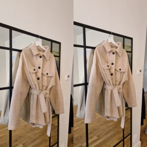 Beige kappa med bälte - Snygg oversize beige kappa med knappar och bälte i midjan. Jackan har långa ärmar och en klassisk krage. Perfekt för höst och vår, med en stilren och tidlös design.