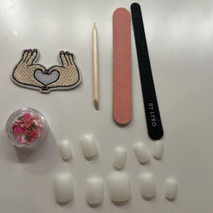 Nagelkit med dekorationer och filar - Komplett nagelkit med vita lösnaglar, två nagelfilar, en träpinne för nagelband och en burk med nageldekorationer. Inkluderar även en söt tygpatch med händer som formar ett hjärta. Perfekt för att skapa en unik nagelstil hemma.