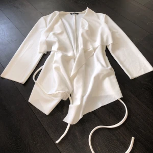 Vit omlottjacka med knytband - Säljer en stilren vit kappa med knytband i midjan. Ganska korta armar, blir mer som trekvarts. Går ungefär till strax under rumpan. Finns en fläck(bild 3) men har man flikarna vid sidan så syns det inte! Använd 1-3 gånger