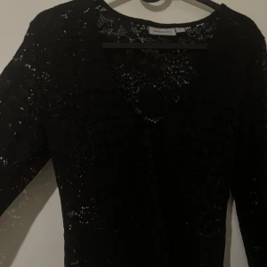 Svart spetsklänning från Kappahl - Elegant svart spetsklänning från Kappahl med v-ringning. Perfekt för en stilfull look. Toppen har ett vackert spetsmönster som ger en sofistikerad touch.