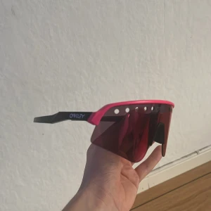 Rosa skidglasögon från Oakley - Snygga skidglasögon från Oakley med en cool rosa båge och mörka linser. Perfekta för utomhusaktiviteter med sin aerodynamiska design och bekväma passform. De har även små ventilationshål för att minska imma. Helt nya, nypris: 2700kr