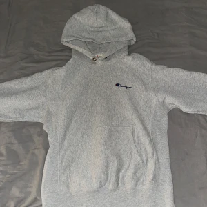 Grå hoodie från Champion - Säljer en klassisk grå hoodie från Champion med broderad logga på bröstet och ärmen. Tröjan har en stor ficka framtill och justerbar huva med snören. Perfekt för en avslappnad stil.