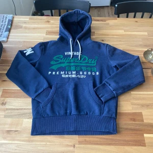 Blå hoodie från Superdry - Säljer en blå hoodie från Superdry med tryck i grönt och vitt på framsidan. Den har en justerbar huva med snören och en stor ficka framtill. Perfekt för en avslappnad stil. Storlek S inga defekter passar M också!