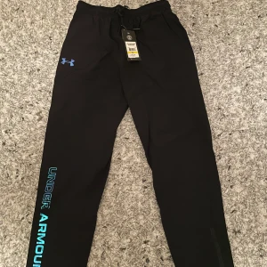 Svarta träningsbyxor från Under Armour - Snygga svarta träningsbyxor från Under Armour med blå logga och text längs benet. Byxorna har en loose passform och är perfekta för träning. De har elastisk midja med snörning för extra komfort. Passar mer som M-L
