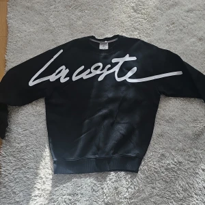 Svart sweatshirt från Lacoste Live - Säljer en svart sweatshirt från Lacoste Live med stor vit logotyp på framsidan. Tröjan är unisex och har en klassisk rund halsringning. Den ikoniska Lacoste-krokodilen är broderad på baksidan av kragen. Perfekt för en stilren och avslappnad look.