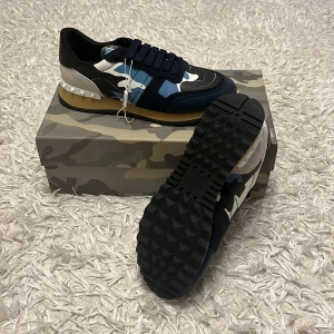 Valentino Rockrunner i blått och kamouflage - Snygga Valentino Rockrunner sneakers med ett coolt kamouflagemönster i blått, vitt och svart. Skorna har en robust gummisula och klassisk snörning. Perfekta för att ge din outfit en unik touch.