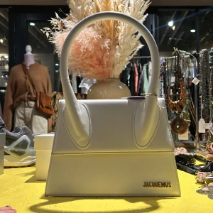 Vit Jacquemus handväska - Snygg vit handväska från Jacquemus med stilren design och rundade handtag. Väskan är i den stora modellen. Väskan har en elegant struktur och är perfekt för att ge din outfit en lyxig touch. Den har en diskret logotyp i guld på framsidan och en praktisk insida för dina nödvändigheter.