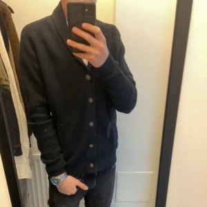 Cardigan - Säljer en stilren mörkblå kofta med knappar framtill. Koftan har en ribbad struktur och långa ärmar, perfekt för kyligare dagar. Den har en klassisk design som passar till många olika outfits. Inga defekter. 