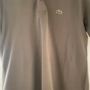 Brun pikétröja från Lacoste - Snygg brun pikétröja från Lacoste med klassisk krage och knappar framtill. Den har korta ärmar och det ikoniska gröna krokodilemblem på bröstet. Perfekt för en stilren och avslappnad look. Tveka inte på att höra av dig vid frågor eller funderingar. Mvh Sixten 