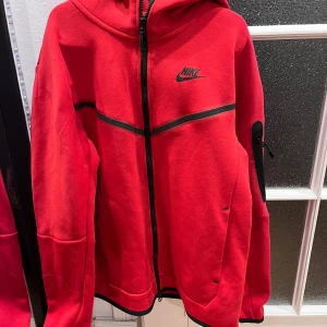Röd huvtröja från Nike - Säljer en röd huvtröja från Nike med dragkedja och svarta detaljer. Tröjan har en justerbar huva och långa ärmar. Perfekt för en sportig look eller en avslappnad dag.