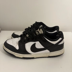 Nike Dunk Low svart/vit sneakers - Svarta och vita Nike Dunk Low sneakers med klassisk svart swoosh på sidan. Skorna har snörning, perforerad tå och platt sula. Stilren färgkombination som passar till det mesta. Ovandelen är i skinn och textil, med lågt skaft och svartvit design.