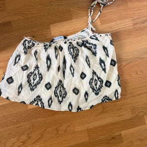 Vit och svart mönstrad linnetopp - Säljer en luftig vit linnetopp med svart grafiskt mönster och smala flätade axelband. Toppen är ärmlös och har en avslappnad passform, perfekt för varma dagar. Tyget är lätt och känns skönt mot huden.